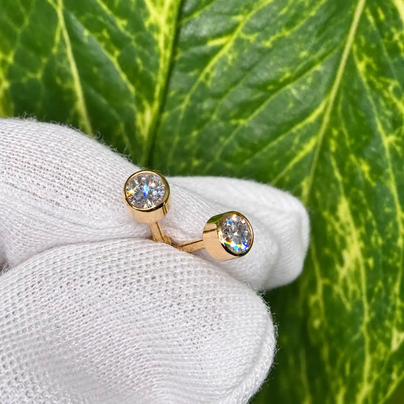 Two 14k yellow gold 4mm diamond bezel-set flat back earrings held in a white-gloved hand against a green leaf