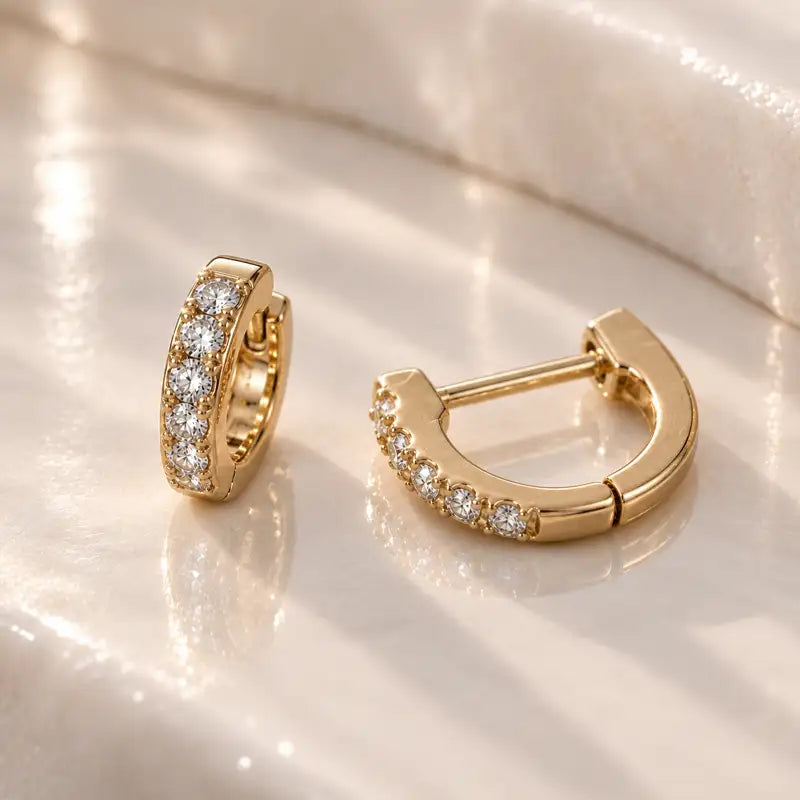 A pair of gold huggie hoop earrings with rows of diamonds, displayed on a reflective white surface.