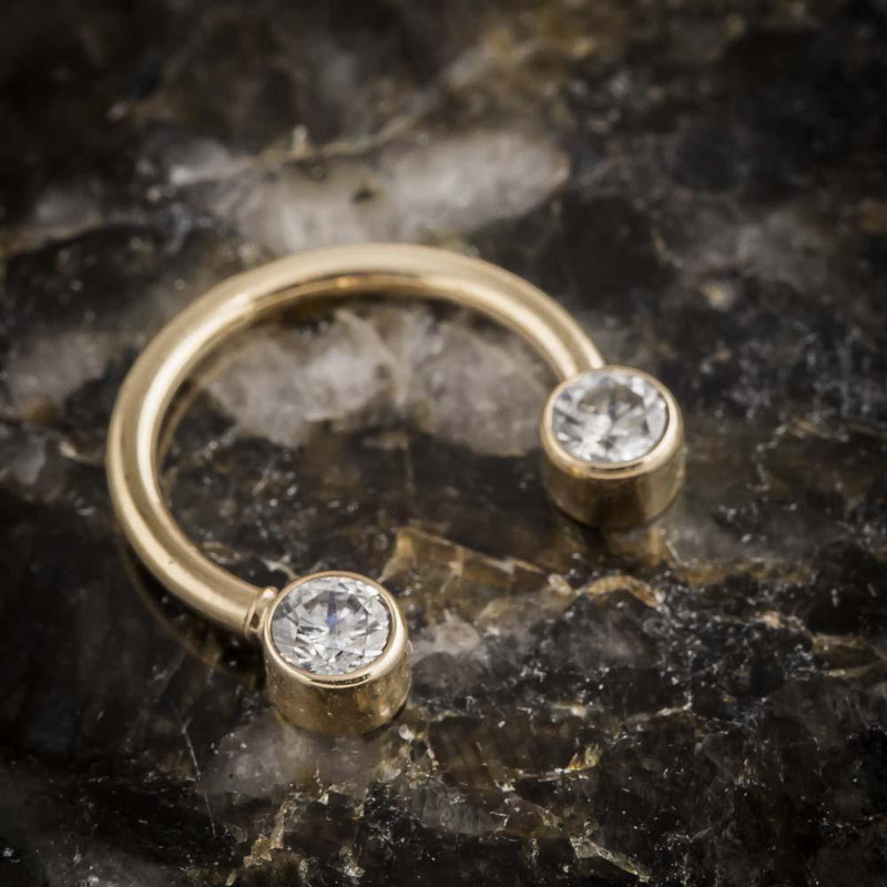 Close-up of 14K yellow gold circular barbell showing bezel-set diamond ends on dark mineral surface
