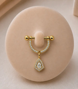 A yellow gold stirrup piercing with pavé-set clear stones and a dangling pear-shaped clear stone pendant, displayed on a round beige jewelry stand.