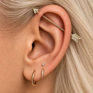 Close-up of a person's ear showcasing multiple gold ear piercings, including an industrial barbell piercing, a small hoop, a pavé stud, and a pavé hoop.