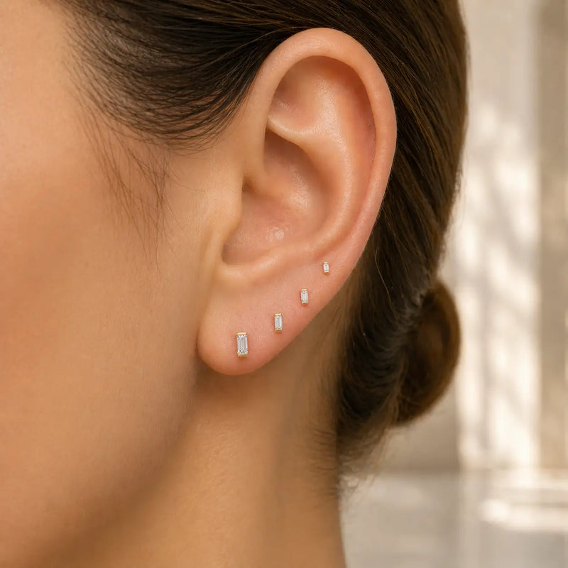 Four baguette diamond push pin studs in graduated sizes worn in stacked lobe piercings, 14k yellow gold