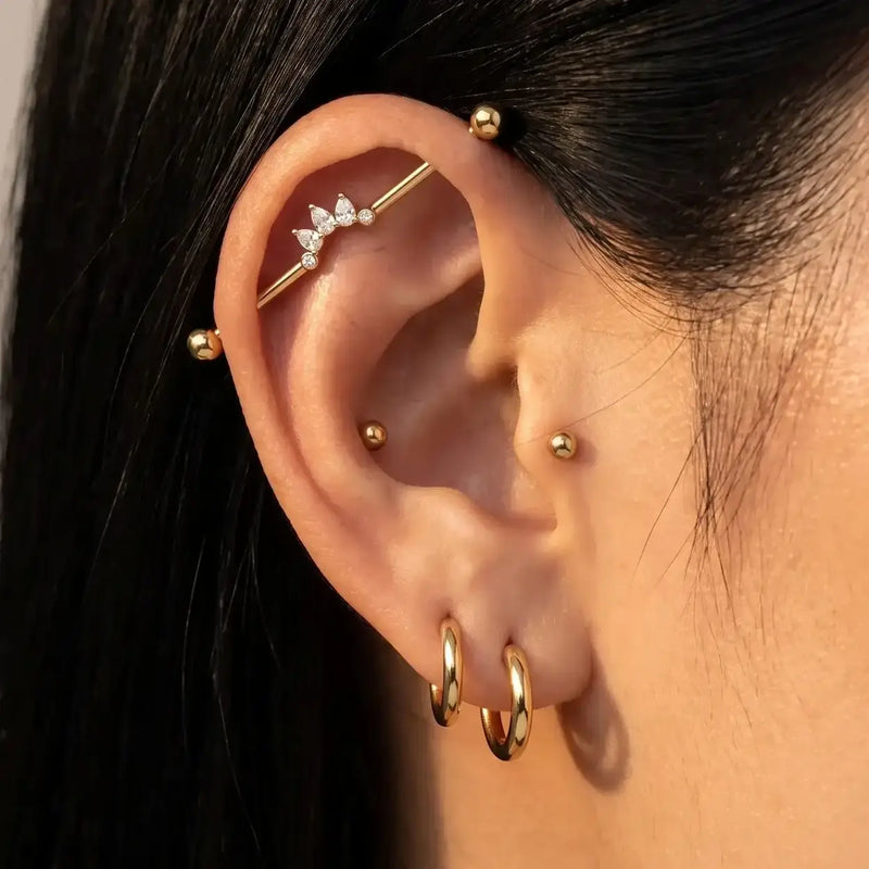 Close-up of a person's ear adorned with multiple gold ear piercings, including hoop earrings, small studs, a helix stud, and the Celeste Arc industrial barbell.