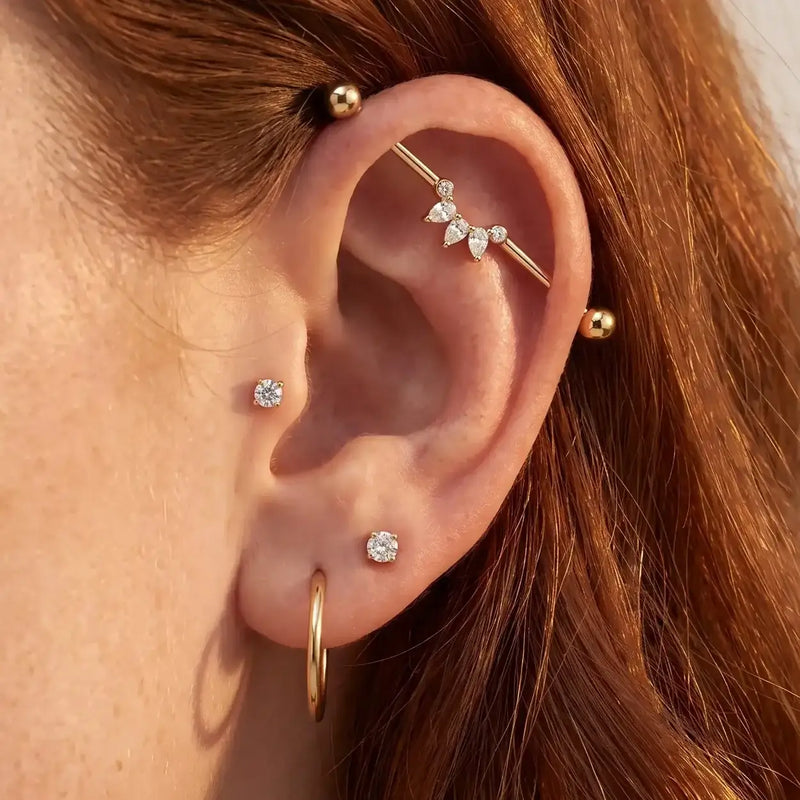 Close-up of a person's ear adorned with multiple gold ear piercings, including hoop earrings, small studs, a helix stud, and the Celeste Arc industrial barbell.