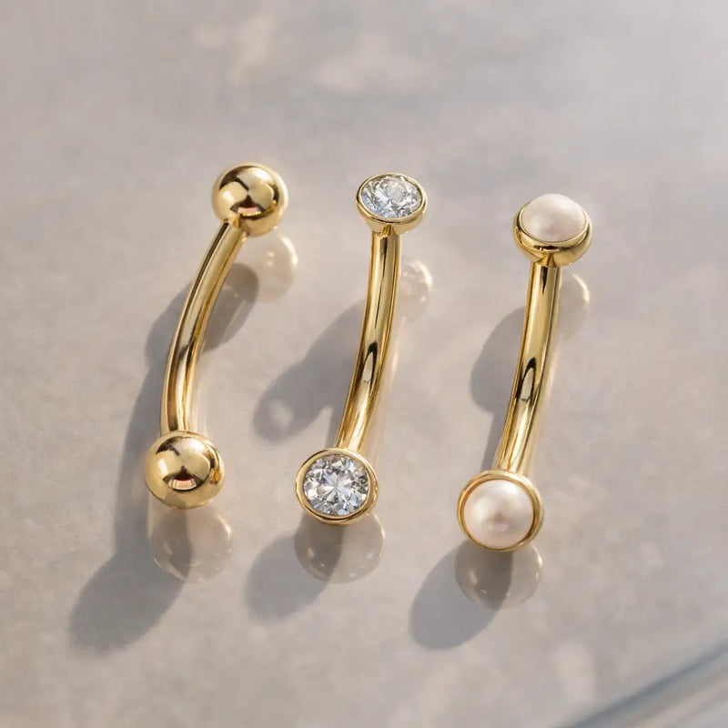 14k yellow gold curved barbells styled alongside CZ and pearl end variations on marble surface