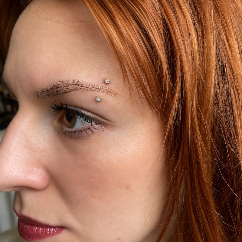 Model wearing a 14k gold prong-set curved barbell with 3mm clear gems in an eyebrow piercing - shown for size and placement; the ruby version is the same size