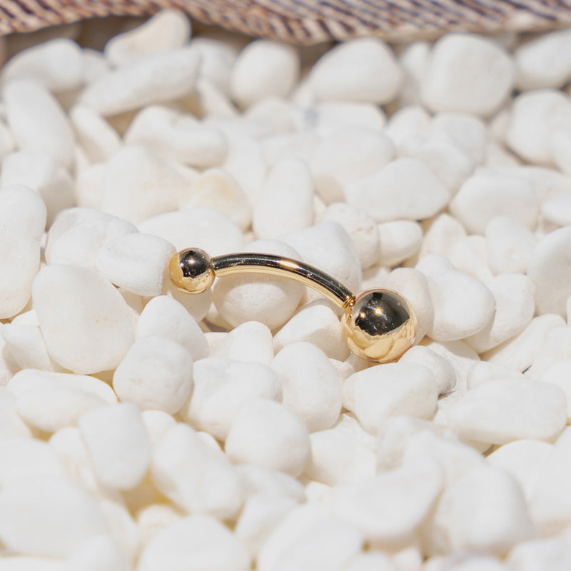 14k yellow gold double ball belly ring resting on white decorative pebbles, angled lifestyle shot