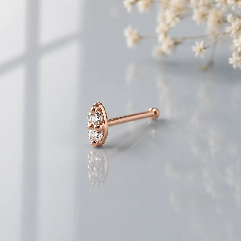 14K rose gold double diamond nose bone style, angled side view on cool gray reflective surface