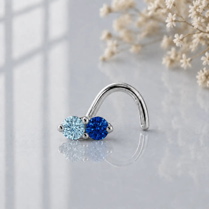 Aquamarine and blue sapphire white gold nose ring on a reflective glass with flowers in the background.