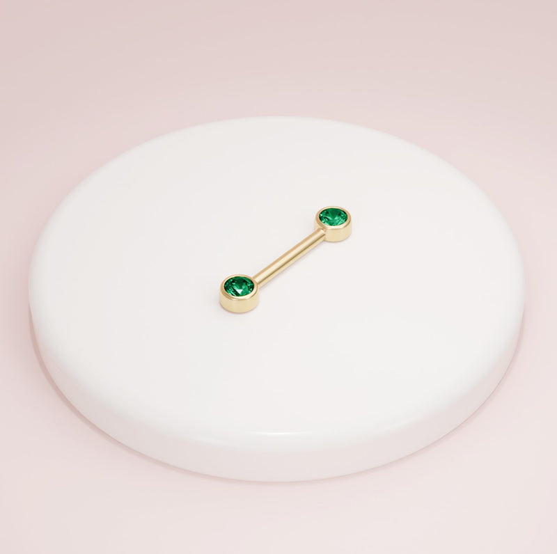 14K yellow gold emerald bezel nipple barbell resting on white ceramic tray, lifestyle shot