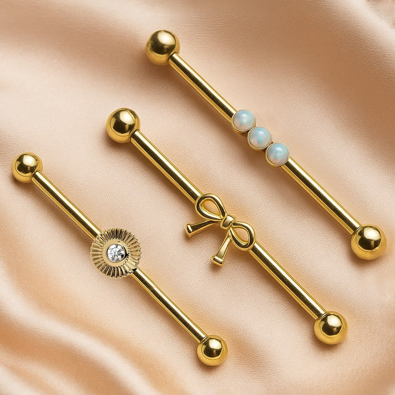 Bow industrial barbell in 14k gold displayed alongside sunburst CZ and opal style barbells on satin