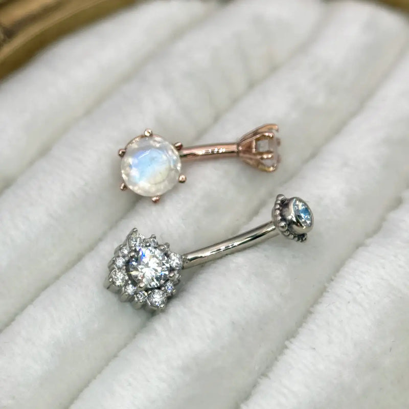 Two belly rings displayed on a white textured ring pad: one rose gold piece with a moonstone, and one white gold piece with diamonds.