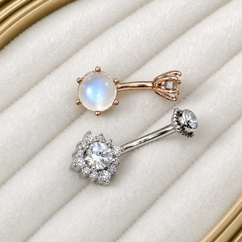 Two belly rings displayed on a white textured ring pad: one rose gold piece with a moonstone, and one white gold piece with diamonds.