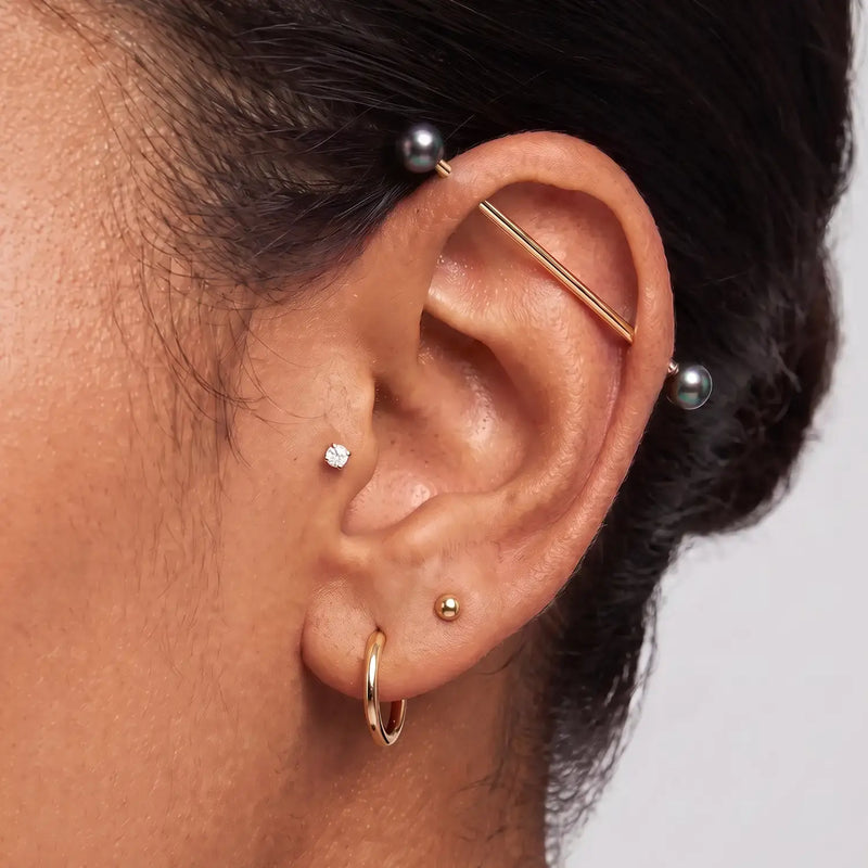 A close-up of a person's ear showcasing multiple earrings, including an industrial barbell with iridescent peacock pearl ends.
