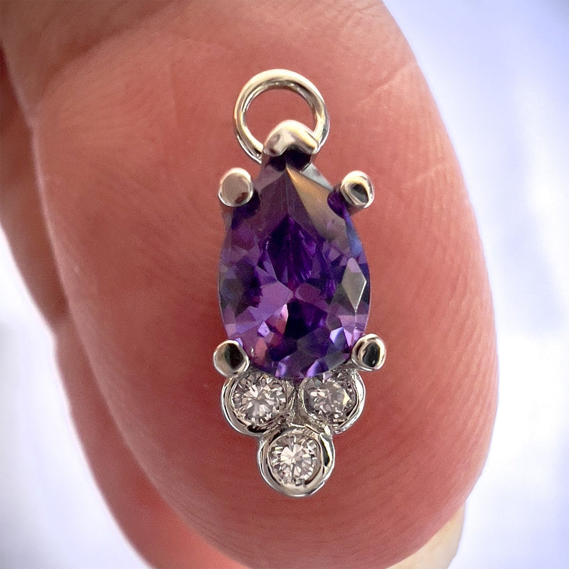 Close-up of amethyst pear charm balanced on fingertip, showing prong setting and diamond trio detail