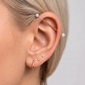 A close-up of a person's ear showcasing a multi-piece gold hoop set, including an industrial barbell with white pearl ends.