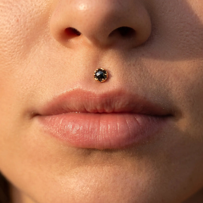 Close-up of a person's nose and lips wearing a small gold stud with a dark round gemstone in the medusa.