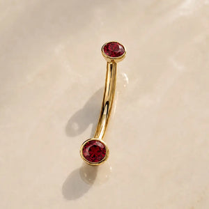 A curved gold barbell with two round faceted ruby gemstones, displayed on a light beige marble surface.