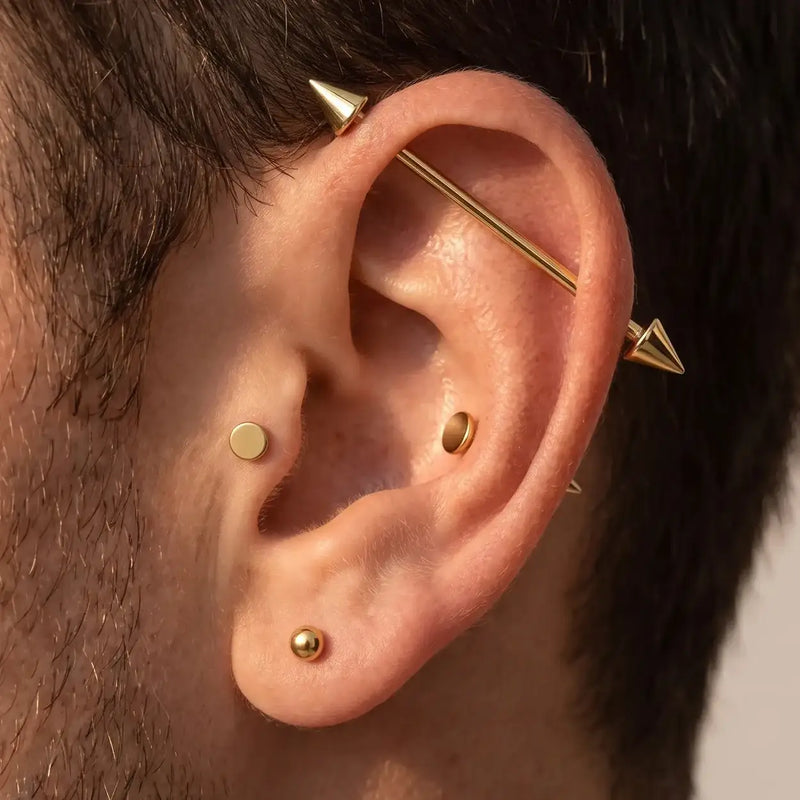 Close-up of a man's ear showcasing multiple gold piercings, including a flat stud, a barbell piercing, and spiked industrial barbell.