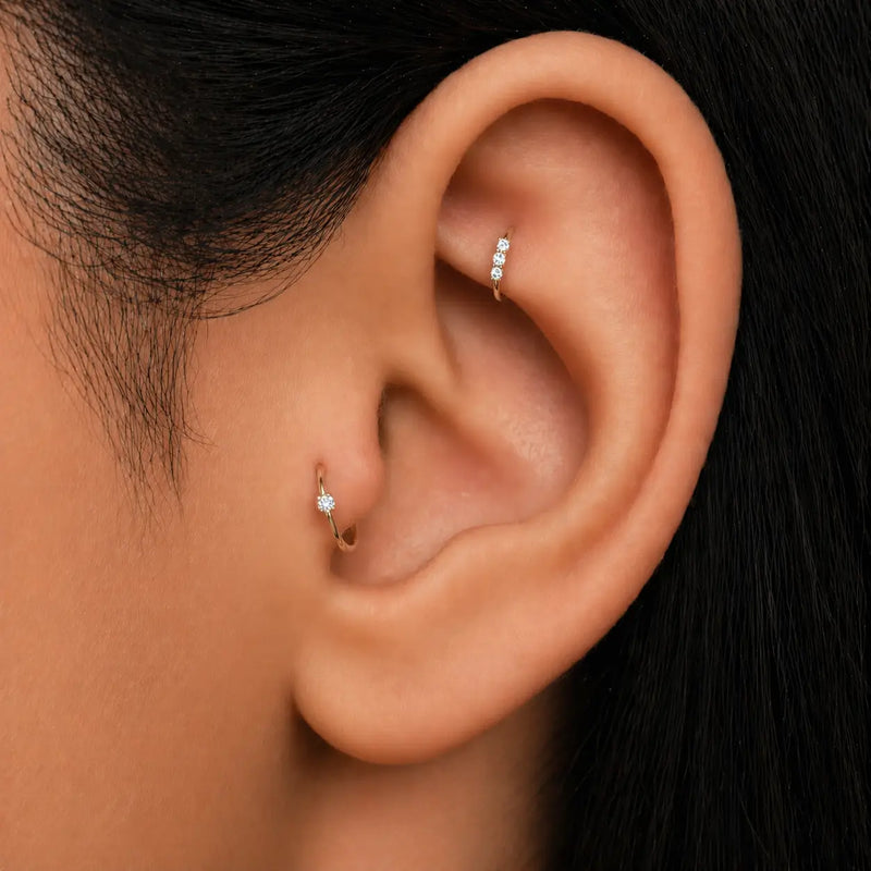Three-diamond gold seam ring worn in helix piercing, shown alongside a tragus hoop