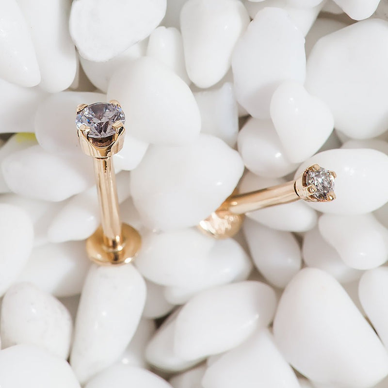 Two 14k rose gold 2mm diamond flat back labret studs resting on white decorative pebbles