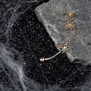 14K rose gold spider curved barbell and matching stud displayed on dark stone with cobweb styling