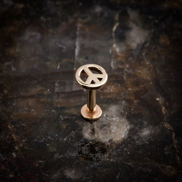 14K rose gold peace sign labret stud standing upright on dark stone surface, full profile view