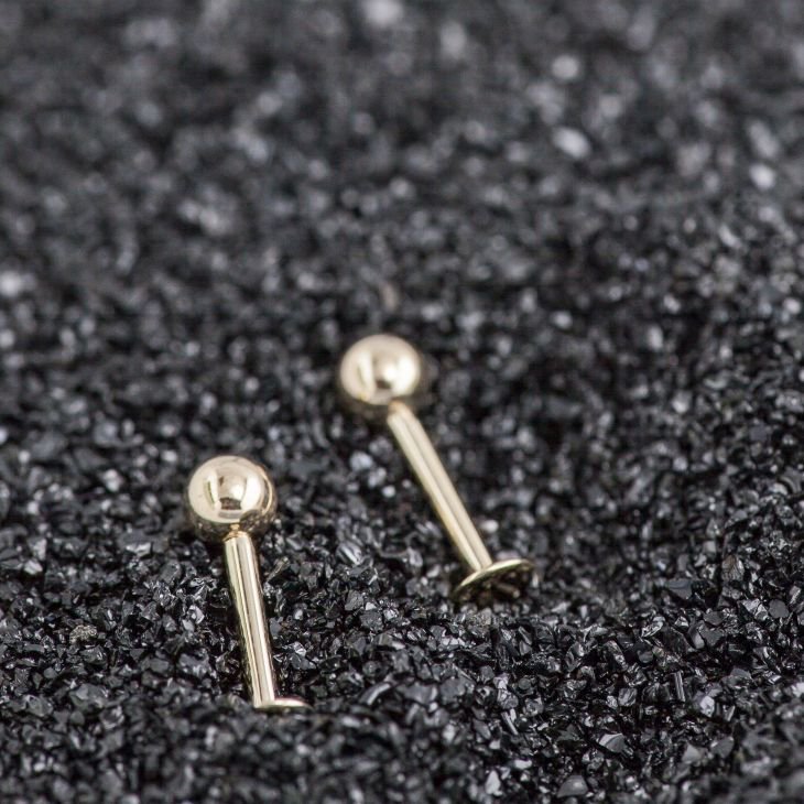 Two 14K yellow gold ball flat back labret studs in different lengths resting on dark textured surface