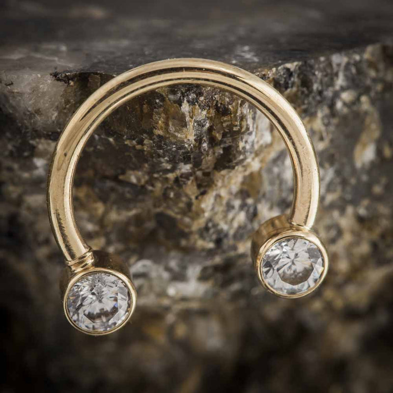 14k yellow gold circular barbell with clear CZ bezel ends resting against rough stone surface