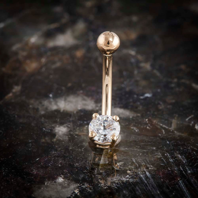 14k rose gold navel ring with round CZ prong setting, upright on dark stone surface