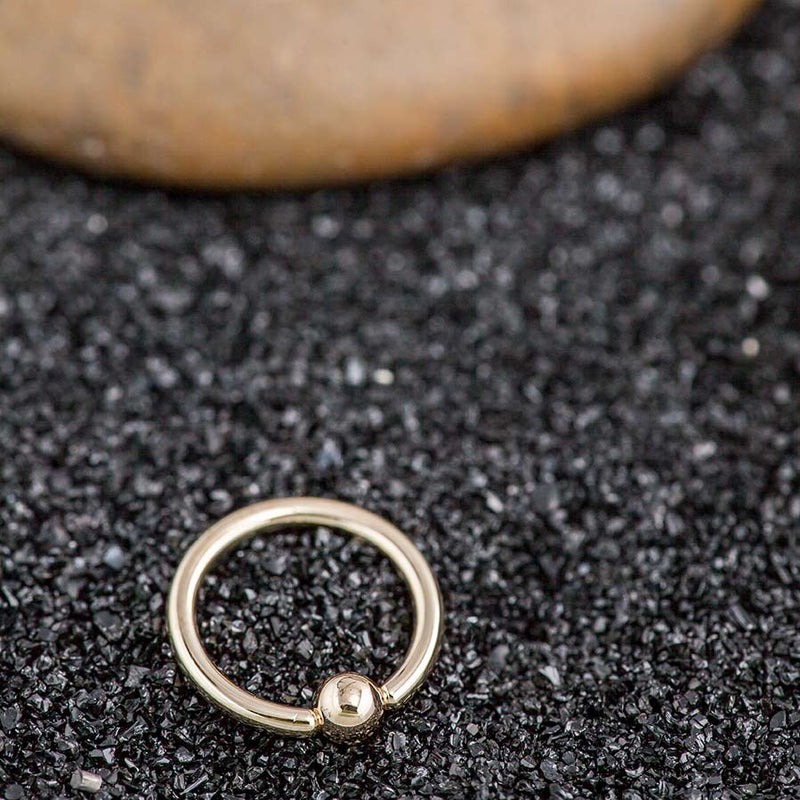 Single 14k gold fixed bead ring on black gravel surface showing hoop and bead closure detail