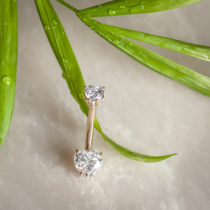 14k yellow gold heart CZ navel ring standing upright against tropical green leaves backdrop