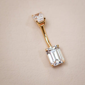 14k yellow gold double emerald-cut clear CZ navel ring laid flat on neutral surface, angled view