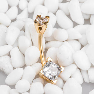 14k yellow gold double princess-cut CZ navel ring resting on white pebbles, angled side view