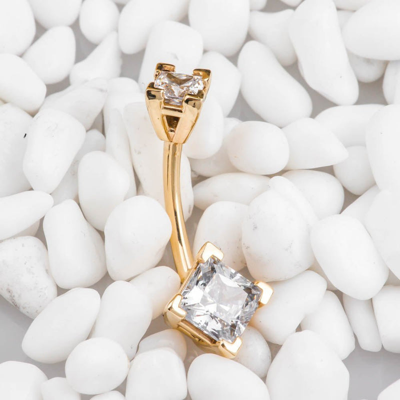 14k yellow gold double princess-cut CZ navel ring resting on white pebbles, angled side view