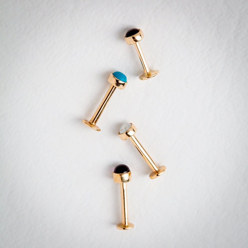Four yellow gold flat back labret studs with cabochon tops laid flat, including opal, turquoise, and onyx options