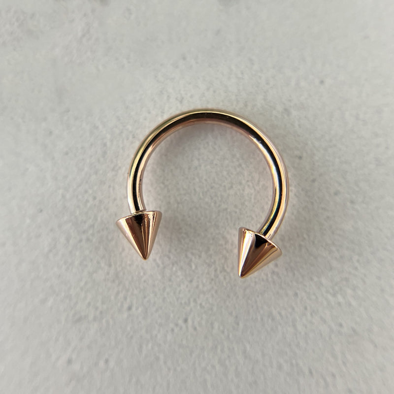 14K rose gold spike horseshoe barbell laid flat on stone surface, top-down view