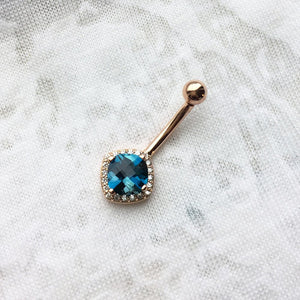 14k rose gold Swiss blue topaz and diamond navel ring laid flat on white fabric, angled view