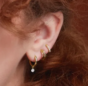 Gold huggie hoop earrings styled on pierced ear alongside ear cuff and charm hoop