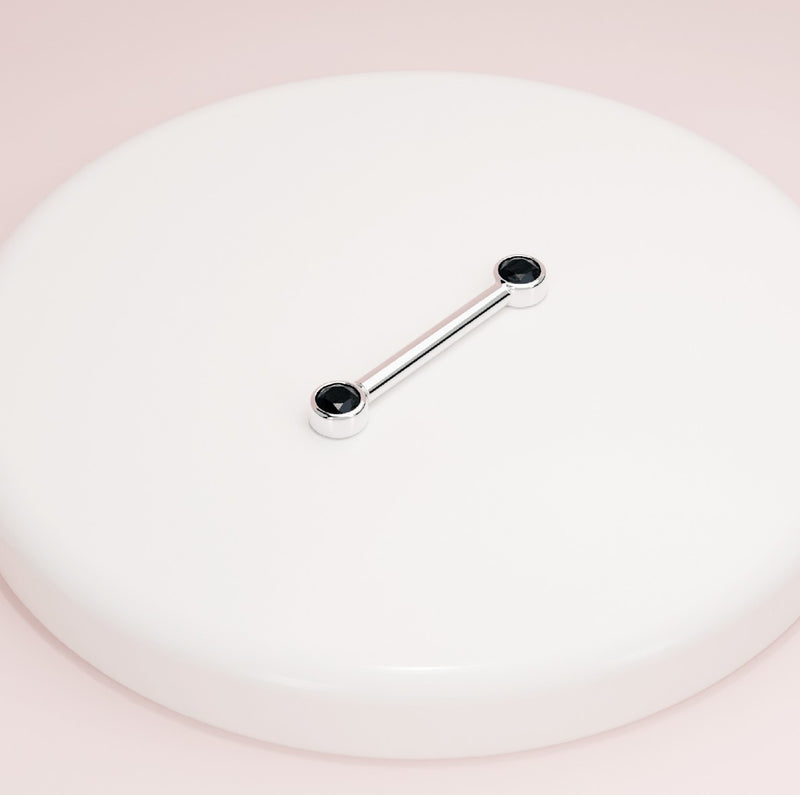 14K white gold black diamond bezel nipple barbell resting on white surface, angled lifestyle shot