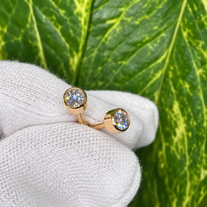 Pair of 14k yellow gold 4mm diamond bezel-set stud earrings held in a white-gloved hand against a green leaf