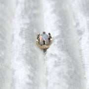 A small gold crown-shaped ring setting holding an iridescent white opal, resting on a textured white fabric background.