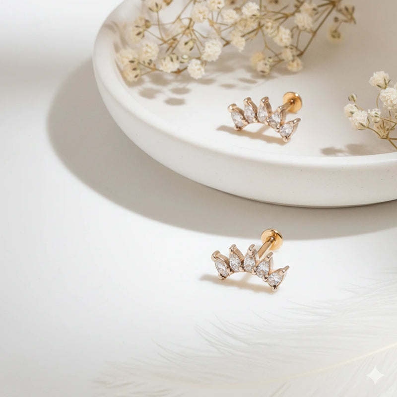 Two 14k rose gold white sapphire crescent cartilage studs displayed on ceramic tray with dried flowers