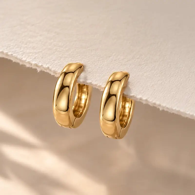 A pair of polished gold huggie hoop earrings resting on a textured white surface with soft shadows.