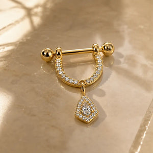 14K yellow gold and diamond-paved nipple stirrup with a dangling teardrop-shaped diamond halo charm, displayed on a beige marble surface with soft shadows.