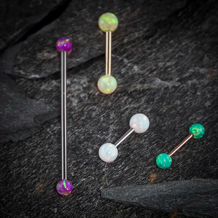 Assorted opal industrial barbells in purple, white, and green on dark stone surface showing color variety