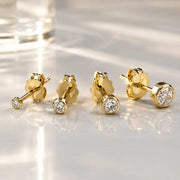 Four 14K yellow gold bezel-set diamond studs in graduated sizes arranged on a reflective surface