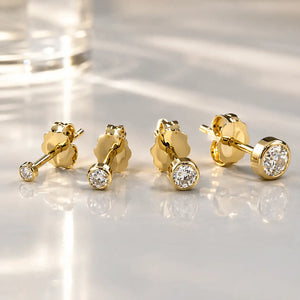 Four 14K yellow gold bezel-set diamond studs in graduated sizes arranged on a reflective surface