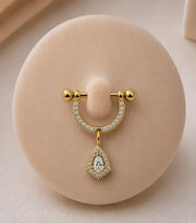 A yellow gold stirrup piercing with pavé-set clear stones and a dangling pear-shaped clear stone pendant, displayed on a round beige jewelry stand.