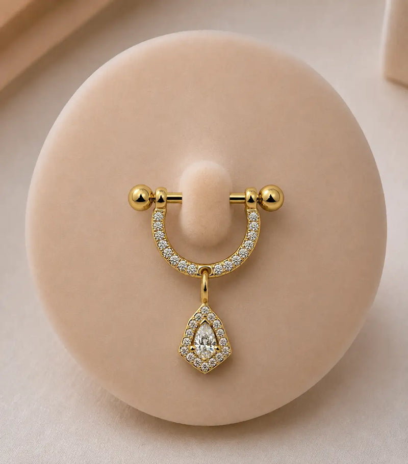 A yellow gold stirrup piercing with pavé-set clear stones and a dangling pear-shaped clear stone pendant, displayed on a round beige jewelry stand.